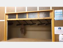 Holzgerahmte Garderobe mit Glasoberteil, mehreren Hakenreihen und einer aufgehängten Jacke. An der rechten Seitenwand sind Aushänge mit Hygienehinweisen und ein Desinfektionsspender sichtbar.