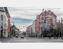 Straßenszene in einer urbanen Umgebung mit mehrstöckigen Wohnhäusern, eines davon mit einer großen, bunten Wandmalerei. Die Straße ist mit Autos, Radfahrern und Fußgängern belebt, über ihr verlaufen zahlreiche Straßenbahnleitungen.