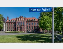 Backsteingebäude im neugotischen Stil mit Spitzbogenfenstern und einem roten Satteldach, umgeben von grünen Bäumen und einer Rasenfläche. Im Vordergrund steht ein Straßenschild mit der Aufschrift „Platz der Freiheit“.