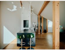 Offene, moderne Wohnküche mit hohen weißen Wänden, Holzstützen und einem dunklen Kochblock mit zwei grünen Barhockern. Die Holzböden und große Fenster sorgen für eine helle und warme Atmosphäre.