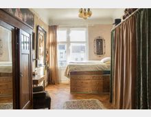 Ein gemütliches Schlafzimmer mit Holzboden und antikem Möbelstil; ein hohes Bett steht auf Holzkästen mit Schubladen, gegenüber ein großer Kleiderschrank mit Spiegel. Die Fenster lassen Tageslicht herein, daneben hängen Vorhänge aus goldfarbenem Stoff.