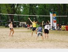 Eine Gruppe von Personen spielt Beachvolleyball auf einem Sandplatz in einem Park mit Bäumen im Hintergrund. Im Hintergrund sind Sitzbereiche, weitere Personen und bunte Banner sichtbar.