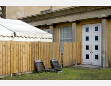 Außenansicht eines Gebäudes mit gelbem Backstein, weißen doppelflügeligen Türen und Fenstern mit Sprossen. Im Vordergrund stehen ein Holzlattenzaun, ein weißes Zeltdach und mehrere zusammengeklappte Stühle auf einer Rasenfläche.