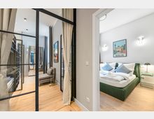 Heller, modern eingerichteter Apartmentflur mit Glas-Metall-Trennwand und bodentiefen Vorhängen, Holzboden und Sitzbereich. Rechts öffnet sich ein Schlafzimmer mit grünem Polsterbett, Nachttischen und einer „Berlin“-Wandgrafik.