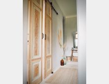 Flur mit hellen Holzboden und Schiebetüren aus naturbelassenem Holz an einer Schiene. Im Hintergrund Dekoration mit getrockneten Gräsern in einer braunen Glasvase sowie ein kleiner Tisch mit Körben.