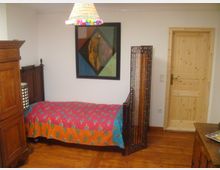 Ein kleines Schlafzimmer mit Holzboden, einem schmalen Bett mit farbenfroher Tagesdecke, einem hölzernen Kleiderschrank, und einer Holztür. An der Wand hängt ein geometrisches Gemälde, und eine bunte Lampe hängt von der Decke.