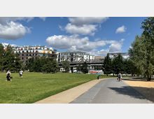Städtischer Park mit einer breiten Wiese, Fußwegen und Bäumen im Vordergrund. Im Hintergrund moderne Wohngebäude mit Balkonen, eine metallene Eisenbahnbrücke und Graffiti an einer Wand.