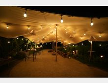 Nachtszene unter einem großen hellen Stoffdach mit warmweißen Lichterketten. Darunter befinden sich hohe Tische mit Dekoration und Sitzgelegenheiten aus Holzpaletten, umgeben von Pflanzen und kleinen Laternen.