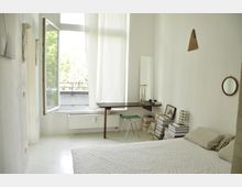 Schlichtes Schlafzimmer mit weiß gestrichenen Wänden, einem Bett im Vordergrund und einem Wandtisch mit Spiegel und Bücherstapeln. Ein großes Fenster mit halb geöffneten Vorhängen lässt Tageslicht in den Raum und zeigt Bäume draußen.