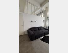 Minimalistisches Loft-Wohnzimmer mit weißer Trennwand und hohen Oberlichtern, Betonboden und hohen Decken. Vor der Wand steht ein großes schwarzes Sofa; rechts ist eine Backsteinwand mit hohen Fenstern und eine angelehnte Leiter zu sehen.
