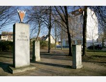 Gedenkstätte in einem Park mit mehreren Steinsäulen, auf der größten steht "Die Toten Mahnen". Umgeben von kahlen Bäumen und angrenzenden Wohnhäusern.