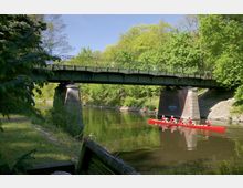 Eine grüne Metallbrücke überspannt einen Fluss, umgeben von dichtem Wald. Im Vordergrund fährt ein rotes Ruderboot mit mehreren Personen auf dem ruhigen Wasser entlang.