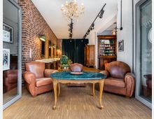 Ein stilvoller Friseursalon mit einer Kombination aus Vintage- und modernen Elementen: Ledersessel, ein grüner Marmortisch, eine Backsteinwand mit Spiegeln und Waschbecken sowie ein Kronleuchter. Im Hintergrund befinden sich ein grüner Samtvorhang, ein antiker Holzschrank und Regale.