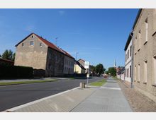 Straßenszene in einem Dorf oder einer Kleinstadt mit gepflastertem Gehweg, einer Fahrbahn und Wohnhäusern mit schlichten Fassaden. Im Hintergrund ist eine Kirche mit einem spitzen Turm und umgebender Vegetation erkennbar.