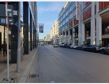 Straßenszene in einer urbanen Umgebung mit mehrstöckigen Büro- und Geschäftsgebäuden auf beiden Seiten. Rechts befindet sich ein Gebäude mit der Beschriftung „Dussmann das KulturKaufhaus“, während geparkte Autos entlang der Straße stehen; im Hintergrund ist eine überdachte Fußgängerbrücke zu sehen.