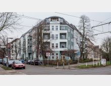 Eckgebäude eines Wohnhauses im Stadtgebiet mit grüner und weißer Fassade, mehreren Balkonen und einem abgerundeten Dachgeschoss. Umgeben von einer Straße mit parkenden Autos, Bäumen und Straßenschildern.
