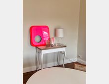 Ein heller Innenraum mit einem weißen Tisch im Vordergrund und einem kleinen weißen Beistelltisch an der Wand. Auf dem Beistelltisch stehen eine weiße Lampe, dekorative Gegenstände und ein großes rotes Dekoelement mit runden Öffnungen. Der Boden ist aus dunklem Holz.