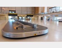 Leerer Gepäckausgabebereich in einer Flughafenhalle mit einem zentralen, ovalen Gepäckband. Im Hintergrund sind hohe Wände, eine Treppe, Leuchtschilder und ein Neon-Restaurant-Schild zu sehen.