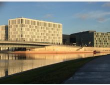 Moderne Bürogebäude in rechteckiger Architektur mit vielen schmalen Fensterreihen, die an einem Flussufer stehen. Im Vordergrund eine Promenade mit Grünfläche und ein Brückenbauwerk, das den Fluss überquert.