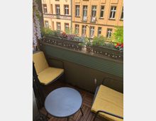 Kleiner Balkon mit Holzboden, zwei gelben Stühlen und einem runden, grauen Tisch. Auf der Brüstung befinden sich zahlreiche Blumentöpfe mit bunten Blumen sowie eine Lichterkette; im Hintergrund sind Altbau-Fassaden mit hellgelbem Anstrich und roten Fensterrahmen zu sehen.