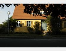 Gelbes Einfamilienhaus mit Satteldach und blauen Fensterläden, umgeben von Bäumen und einem kleinen Vorgarten; eine Straßenlaterne steht links im Bild. Der Eingangsbereich hat eine halbhohe Treppe, die zu einer dunkelblauen Tür führt.