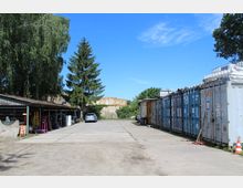 Ein Lagerplatz mit einer Betonfläche, auf der linken Seite eine Holzhalle mit verschiedenen Gegenständen und rechts nebeneinander aufgereihte Frachtcontainer. Im Hintergrund sind ein großer Baum und die Überreste einer alten Ziegelruine sichtbar.