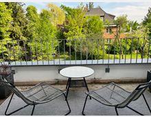 Eine Terrasse mit zwei geflochtenen Stühlen und einem runden weißen Tisch, umgeben von einem Geländer mit Blick auf einen grünen Garten mit Bäumen und einem Backsteinhaus im Hintergrund.