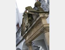 Steinerner Torbogen mit dekorativen Säulen und Verzierungen, auf dem ein Löwenrelief und ein Wappenschild angebracht sind, im Hintergrund eine weiße Fassade mit Bögen und roten Fensterrahmen.