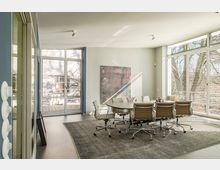 Heller Konferenzraum mit großem runden Tisch und mehreren hellbraunen Bürostühlen auf einem grauen Teppich. Große Fensterfronten mit Jalousien lassen Tageslicht hinein, und im Hintergrund ist eine Außenterrasse zu sehen.