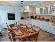 Wohnküche mit weißen Einbauschränken, Arbeitsplatten aus Holz und einer beige gefliesten Rückwand. In der Mitte steht ein Holzesstisch mit sechs Stühlen, über dem eine Hängelampe angebracht ist.