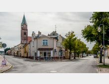 Eine Straßenszene in einer Kleinstadt mit einem beige gestrichenen Eckhaus im Vordergrund, neben dem mehrere Fahrräder geparkt sind. Im Hintergrund ist eine rotbraune Backsteinkirche mit einem hohen Turm und einer Uhr zu sehen, während Bäume entlang der Straße stehen.