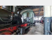 Historische Dampflokomotive und grüner Waggon in einer Scheune oder einem Lokschuppen mit Holzdecke und großen Fenstern. Im Hintergrund ist eine weitere Dampflokomotive zu sehen.