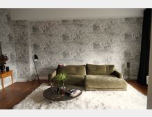 Wohnzimmer mit einer olivgrünen Sofa-Eckkombination vor einer grauen Tapete mit Wolkenmuster. Ein runder Couchtisch mit Dekorationen steht auf einem weißen Teppich, daneben eine Stehlampe, ein kleiner Holztisch und eine schwarze Gardine.