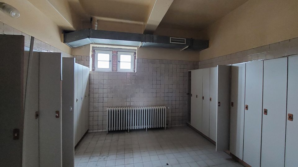 Ein Umkleideraum mit weißen Fliesenwänden, einer Reihe von weißen Spinden auf beiden Seiten, einem Heizkörper unter einem kleinen Fenster und sichtbaren Belüftungsrohren an der Decke. Der Boden besteht aus grauen Fliesen.