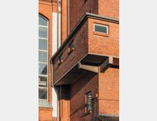 Ziegelsteinfassade eines Gebäudes mit einem Betonbalkon, kleinen rechteckigen Fenstern und fallenden Regenrinnen. Links ist ein großer Fensterbereich mit weißem Rahmen sichtbar.