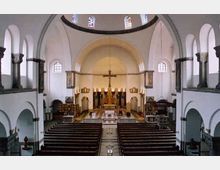 Innenraum einer Kirche mit gewölbter Decke, mehreren Rundbogenfenstern und einem zentralen Altar im Hintergrund. Die Sitzbänke sind symmetrisch angeordnet, und der Raum ist von weißen Wänden sowie dezenten architektonischen Verzierungen geprägt.