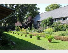 Großer Garten mit gepflegtem Rasen, einer Reihe von Topfpflanzen und bunten Blumenbeeten, umgeben von dichten Bäumen und einem großen Gebäude mit Ziegelfassade und schrägem Dach im Hintergrund.