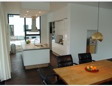 Moderne Wohnküche mit einer zentralen Kücheninsel, integrierten weißen Hochglanzschränken und dunklem Holzboden. Neben einem Holztisch mit schwarzen Lederstühlen hängt eine goldene Hängeleuchte, und durch große Fenster fällt Tageslicht herein.