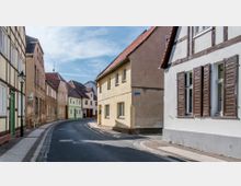 Schmale, gepflasterte Straße in einer Altstadt mit historischen Wohnhäusern; die Fassaden sind teils verputzt, teils aus Fachwerk, mit roten Ziegeldächern und Fensterläden. An der Straße befinden sich Bürgersteige und eine Straßenlaterne.