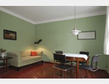 Wohnraum mit grünen Wänden, einer beigen Couch mit einem roten Kissen, einem ausziehbaren Wandlampenarm, einem hölzernen Esstisch mit vier schwarzen Stühlen und hängender Deckenlampe. An den Wänden hängen zwei gerahmte Bilder, und der Boden ist aus glänzendem Holz.