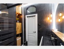 Modernes Badezimmer mit dunklen Wandfliesen, einer weißen Tür mit Oberlicht und Holzakzenten. An der Wand befinden sich offene Hängelampen, ein Holzwaschbeckenregal und ein schmaler schwarzer Waschtisch mit Armaturen.