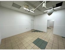 Ein leerer, rechteckiger Raum mit weißen Wänden, halbhoch gefliest, und einem beigefarbenen Fliesenboden mit grauen Akzenten. An der Decke befinden sich ein strahlerähnliches Gerät, Lampen sowie Lüftungsgitter, und es sind zwei Türen sowie ein Durchgang sichtbar.