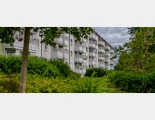 Mehrstöckiges Wohngebäude mit weißen Balkonen, umgeben von dichter grüner Vegetation und Bäumen; der Himmel ist bewölkt.