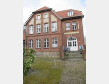 Backstein-Gebäude mit roten Dachziegeln im Stil eines historischen Wohn- oder Schulgebäudes, mit großen Fenstern und einem zentralen Eingang mit weißen Doppeltüren, erreichbar über eine kurze Steintreppe. Im Vordergrund befindet sich ein leicht geneigter Garten mit grasbewachsenem Boden und jungen Bäumen.