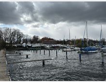 Ein Yachthafen mit Bootsanlegern und festgemachten Segelbooten, einige davon mit Planen abgedeckt. Im Hintergrund stehen Bäume ohne Laub und ein großes Gebäude mit rotem Dach unter einem bewölkten Himmel.