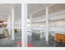 Heller, moderner Innenraum eines Gebäudes mit hohen weißen Säulen, mehreren offenen Etagenebenen und einem breiten Treppenaufgang. Der Boden besteht aus glänzendem dunklem Stein, und an einer Wand sind Holzschließfächer sichtbar.