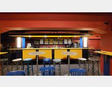 Ein Innenraum einer Bar mit einer gelb-blauen Theke, Regalen mit Gläsern und Flaschen im Hintergrund sowie mehreren hohen Tischen und Stühlen aus Metall im Vordergrund. Die Wände sind in Rot gehalten, und der Boden hat ein dunkles Teppichmuster.