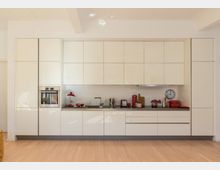 Moderne Einbauküche mit weißen, glatten Schrankfronten, integrierten Geräten und einer langen Arbeitsfläche; hervorgehoben durch Küchenutensilien, wie eine rote Lampe, Toaster und Kaffeemaschine. Holzboden und neutrale Wandgestaltung vervollständigen den minimalistischen Raum.