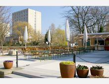 Ein Außenbereich eines Cafés oder Restaurants mit mehreren gruppierten Holzbänken und -tischen sowie geschlossenen Sonnenschirmen; im Hintergrund stehen Laubbäume und ein mehrstöckiges Wohn- oder Bürogebäude. Im Vordergrund befinden sich Blumentöpfe mit bunten Blumen.