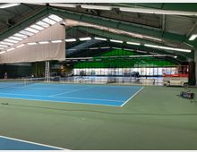 Innenansicht einer Tennishalle mit grünen und blauen Spielfeldern, einer hohen Decke mit Stahlträgern und Dachfenstern. Im Hintergrund sind große Fensterfronten, eine Hebebühne sowie weitere Geräte und Tische sichtbar.